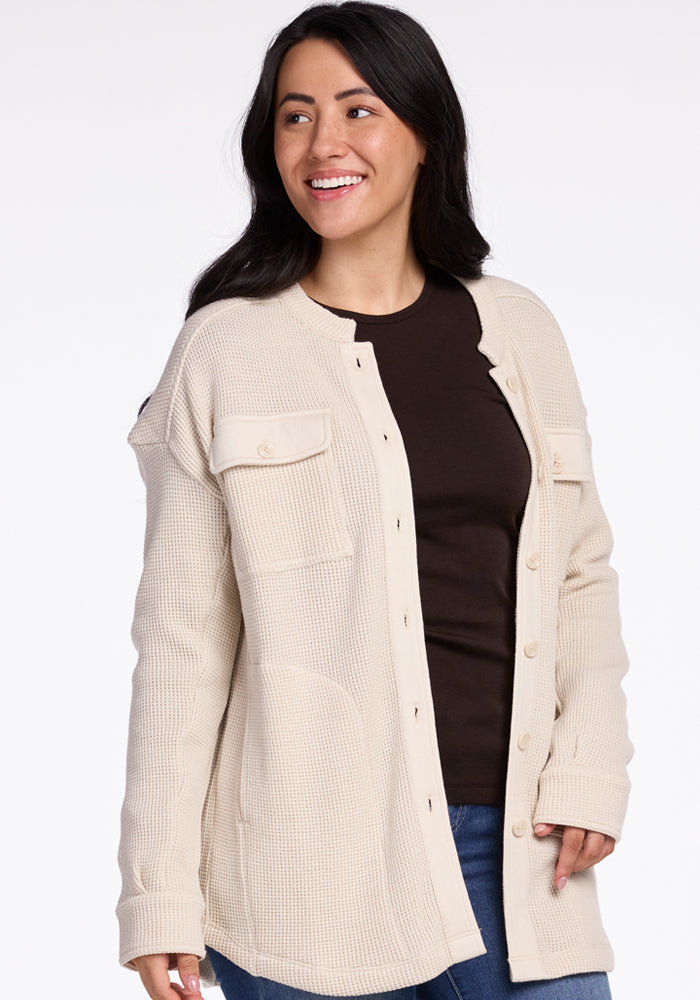 A person smiling, dressed in the Oaklynn Waffle Shacket - Oatmeal by Woolx, layered over a dark shirt and jeans. Crafted from moisture-wicking merino wool, this shacket highlights two chest pockets and long sleeves. They have long dark hair and are positioned against a plain background.