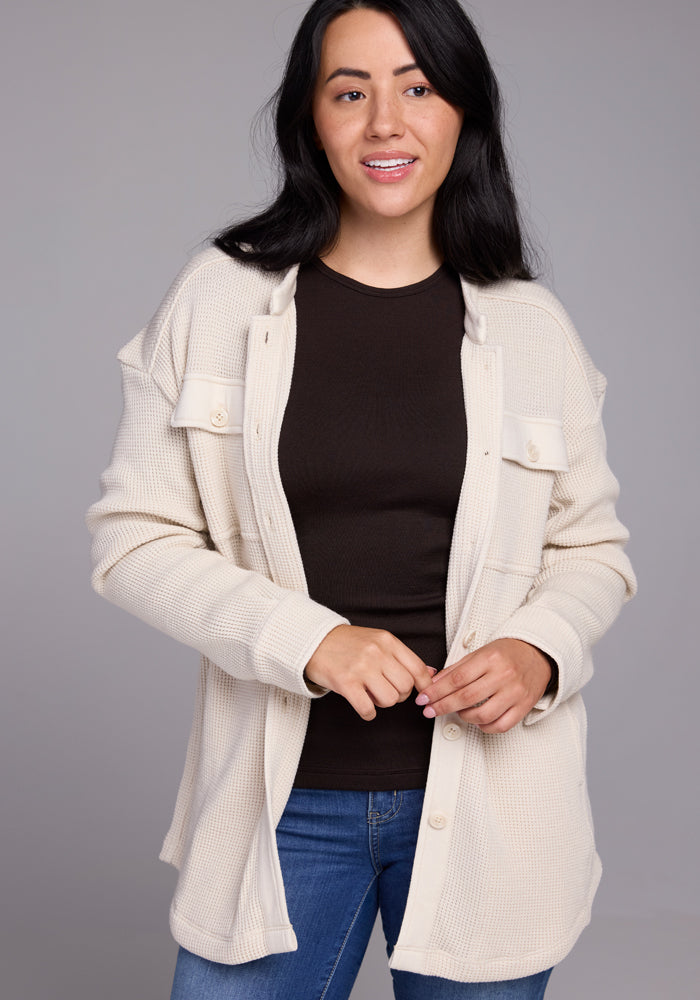 A person with long dark hair is smiling and wearing a black shirt under the Woolx Oaklynn Waffle Shacket in oatmeal. They are standing against a gray background.