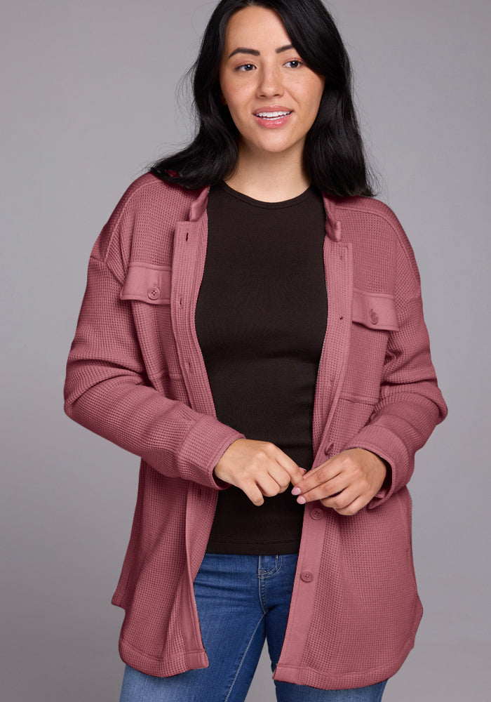 A woman with long dark hair is wearing a black top, blue jeans, and an open Oaklynn Waffle Shacket in Wild Ginger from Woolx. She is smiling slightly, standing against a plain gray background.