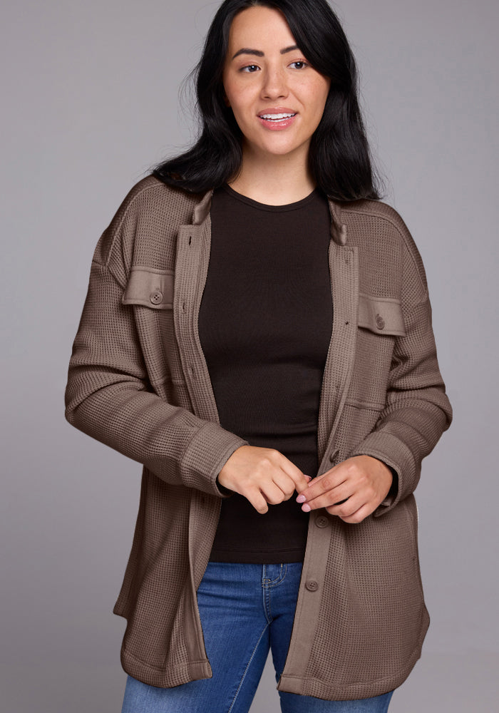 A woman with long dark hair is smiling while wearing the Oaklynn Waffle Shacket in Latte by Woolx. She's standing against a plain grey background, exuding warmth and style.