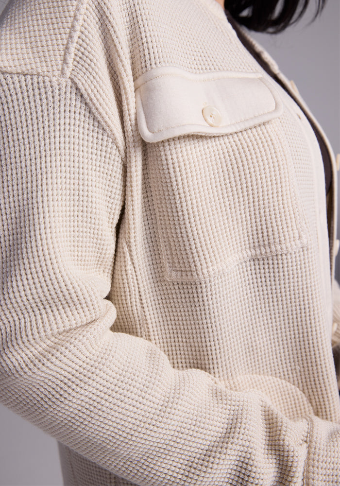 Close-up of a person wearing the Woolx Oaklynn Waffle Shacket in oatmeal, featuring pocket and button detail on the chest. The focus is on the shacket's moisture-wicking texture and the upper body, set against a neutral gray background.