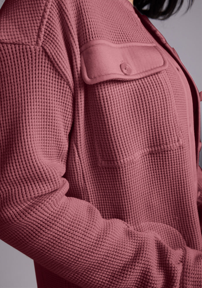 A close-up features someone wearing the Oaklynn Waffle Shacket in Wild Ginger by Woolx, showcasing a textured, buttoned pocket on the chest. The Merino Wool fabric appears soft and comfortable, with the person's hair partially visible.