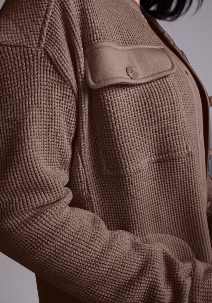 Close-up of a person wearing a textured brown Oaklynn Waffle Shacket - Latte from Woolx, featuring a buttoned chest pocket. The garment has a grid pattern, is slightly oversized, and crafted from moisture-wicking fabric. The neutral background accentuates the texture and details of the shacket.