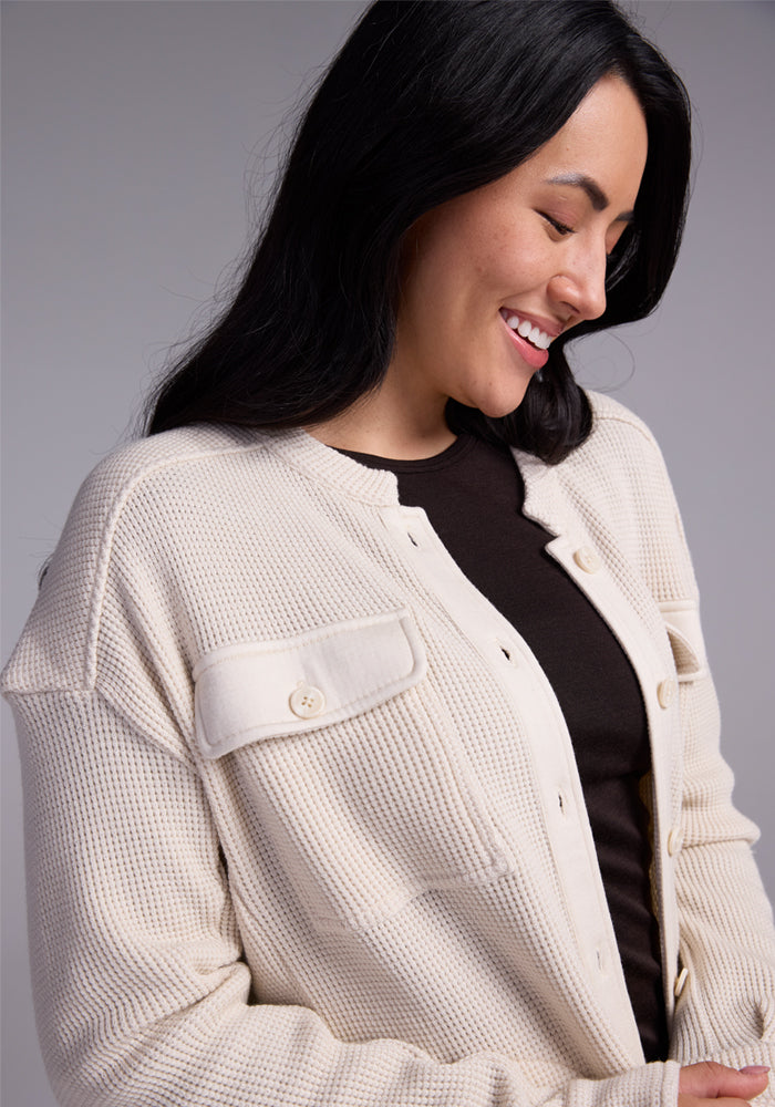 A person with long dark hair smiles while looking down, wrapped in the Woolx Oaklynn Waffle Shacket in oatmeal over a black top. The image captures a relaxed and content expression, with the cozy layer adding an element of warmth and comfort.