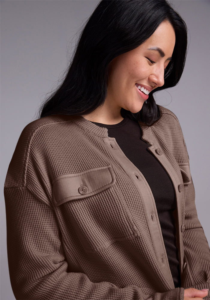 A person with long black hair smiles while looking down, wrapped in the cozy Oaklynn Waffle Shacket - Latte from Woolx over a black top. The brown textured jacket complements the neutral gray background effortlessly.