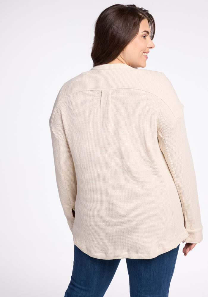 A woman with long dark hair is smiling while looking to her left, dressed in the Oaklynn Waffle Shacket - Oatmeal by Woolx and blue jeans. The photo captures her from the back against a plain white background, emphasizing the texture of her stylish ensemble.