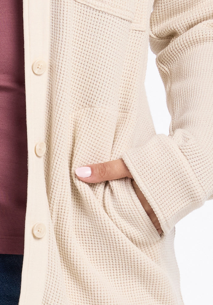 Close-up of a person wearing the Oaklynn Waffle Shacket in oatmeal by Woolx over a maroon top. The shacket has large buttons and a pocket, with the person's hand partially tucked inside, their nails painted light pink.