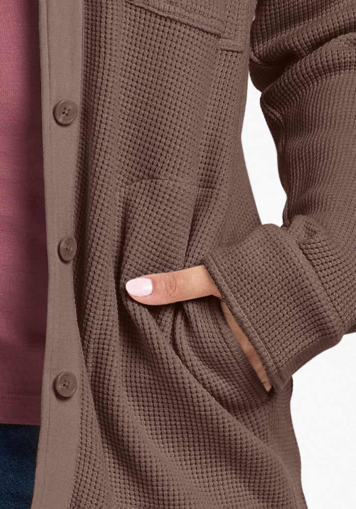 Close-up of a person comfortably wearing a Woolx Oaklynn Waffle Shacket in warm latte, complete with buttons. Their left hand is casually tucked into the pocket, revealing a hint of pink shirt underneath. Paired with dark pants, this Merino wool piece offers both style and comfort for cooler days.