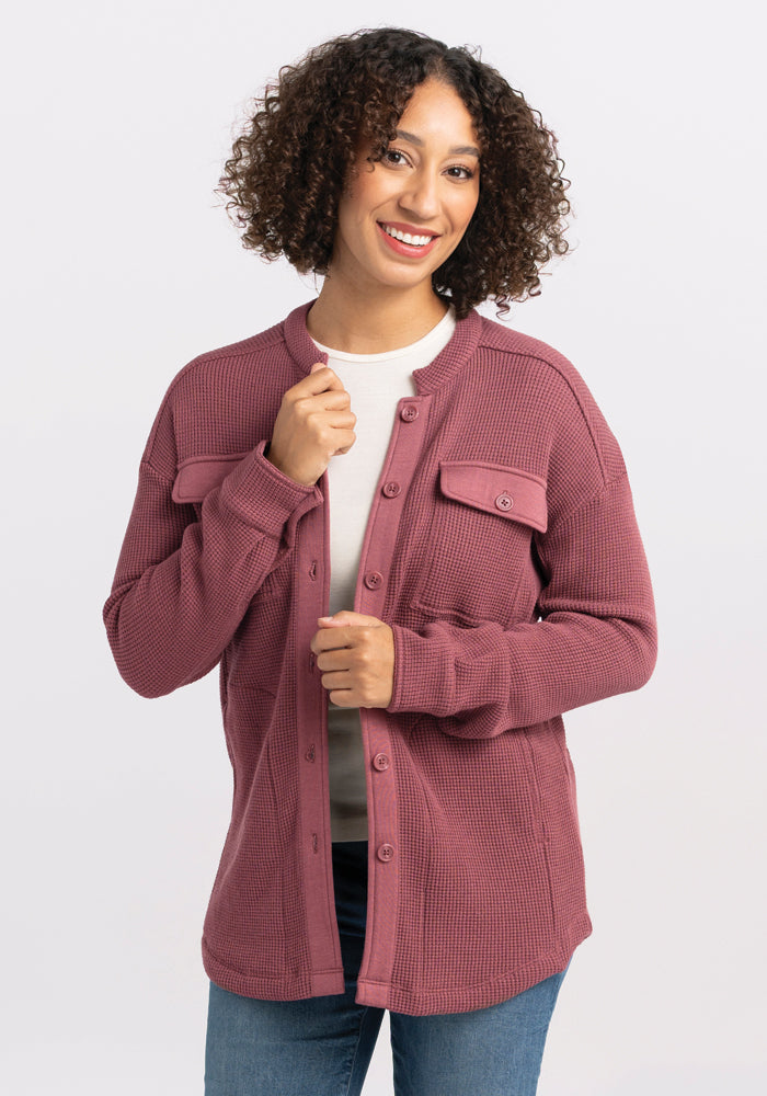 A person with curly hair smiles while wearing an Oaklynn Waffle Shacket in Wild Ginger from Woolx over a white shirt and blue jeans. They stand against a plain white background. 