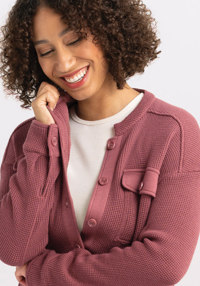 A woman with curly hair, a serene smile on her face and eyes gently closed, is enveloped in the comforting embrace of the Oaklynn Waffle Shacket in Wild Ginger by Woolx, worn over a crisp white shirt. She softly rests one hand against her cheek against a plain white backdrop.