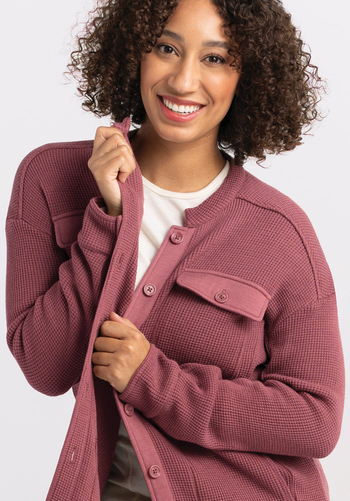 With a warm smile, a person with curly hair is wrapped in the Woolx Oaklynn Waffle Shacket in Wild Ginger. The shacket's textured design and buttoned pockets add depth, as they pull its edge close to their chin.