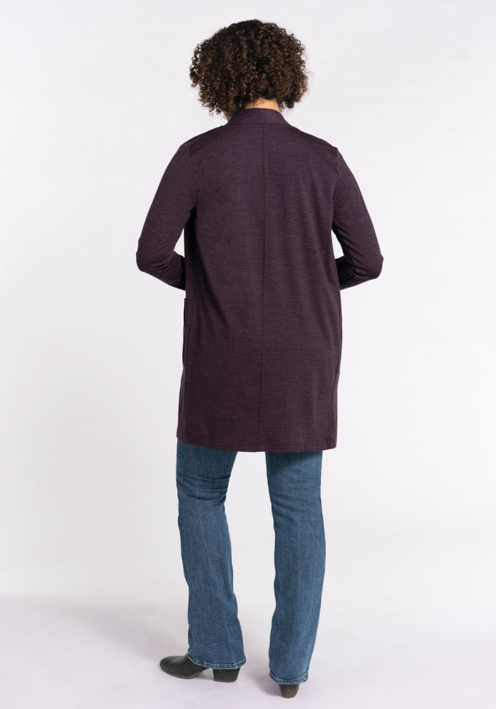 A person with curly hair stands with their back to the camera, wearing a long-sleeved Paisley Cardigan in Deep Plum by Woolx, crafted from luxurious Australian Merino wool. They pair it with blue jeans and black shoes against a plain white background.