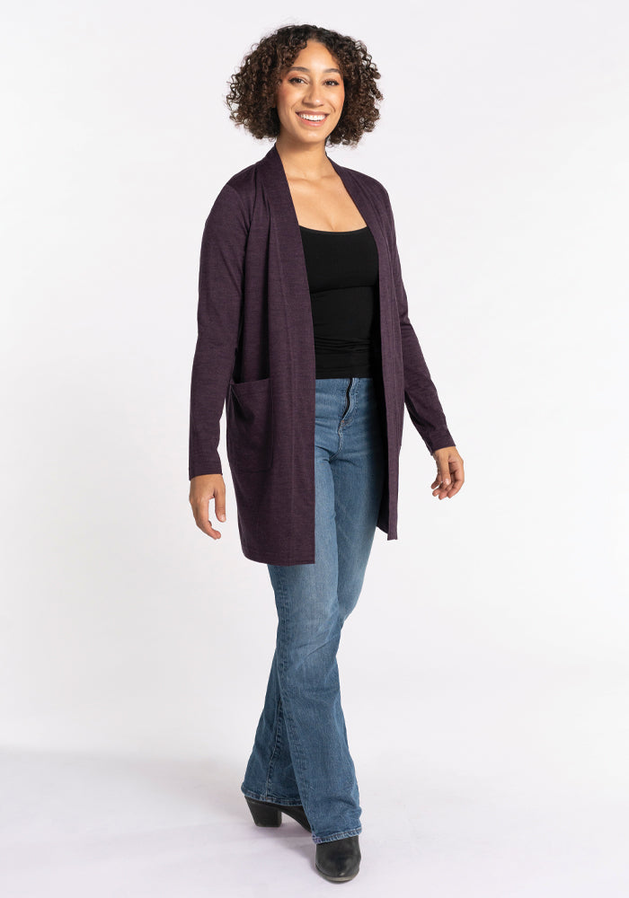 A person with curly hair is smiling, wearing a Woolx Paisley Cardigan in Deep Plum, crafted from Australian Merino Wool, over a black top and blue jeans. They are facing forward with one hand in a pocket, standing on a white background.