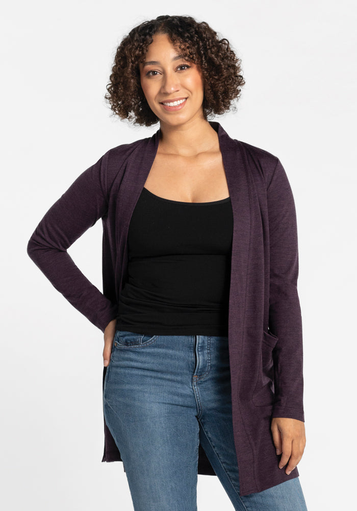 A person with curly hair smiles and stands with one hand on their hip, wearing a Woolx Paisley Cardigan in Deep Plum, crafted from Australian Merino wool. Underneath the cardigan, they have a black top paired with blue jeans, all set against a plain white background.