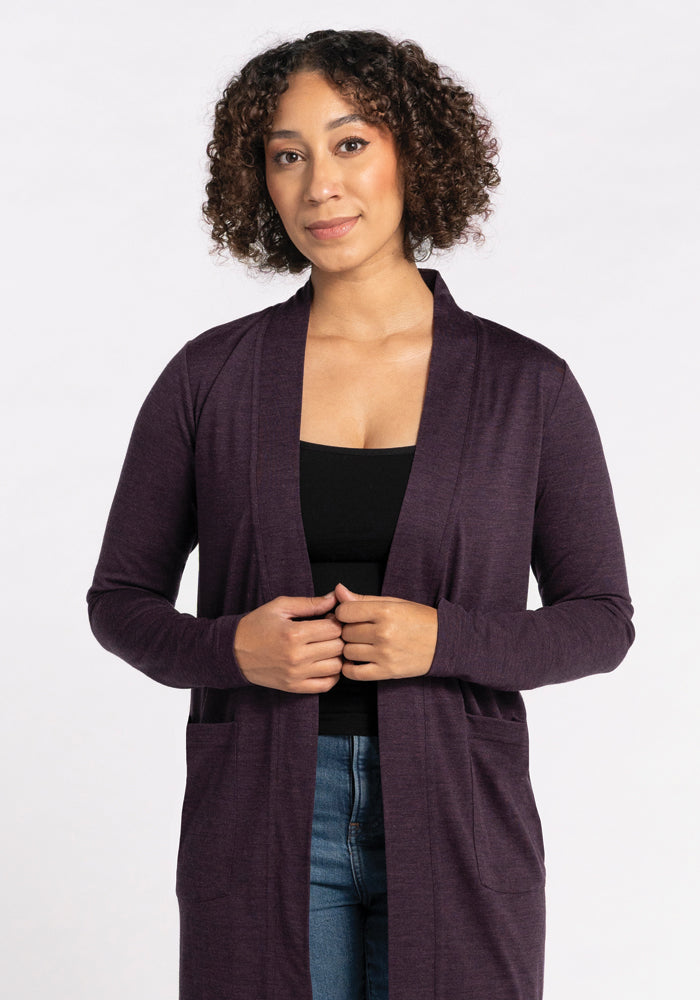 A person with curly hair is wearing a black top and a long Paisley Cardigan in Deep Plum, crafted from Australian Merino Wool by Woolx. They stand against a plain white background, looking forward with a neutral expression and hands gently clasped.