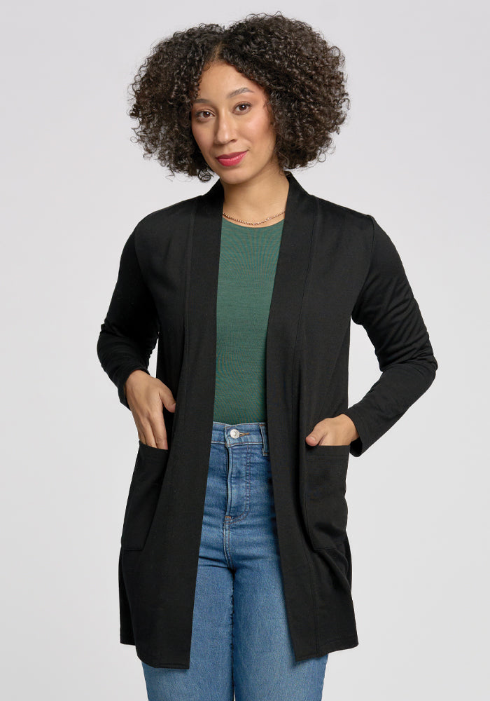 A curly-haired person wears a Woolx Paisley Cardigan in black over a green top and blue jeans. Made from lightweight Merino wool, they stand with hands in the cardigan pockets against a plain background. 