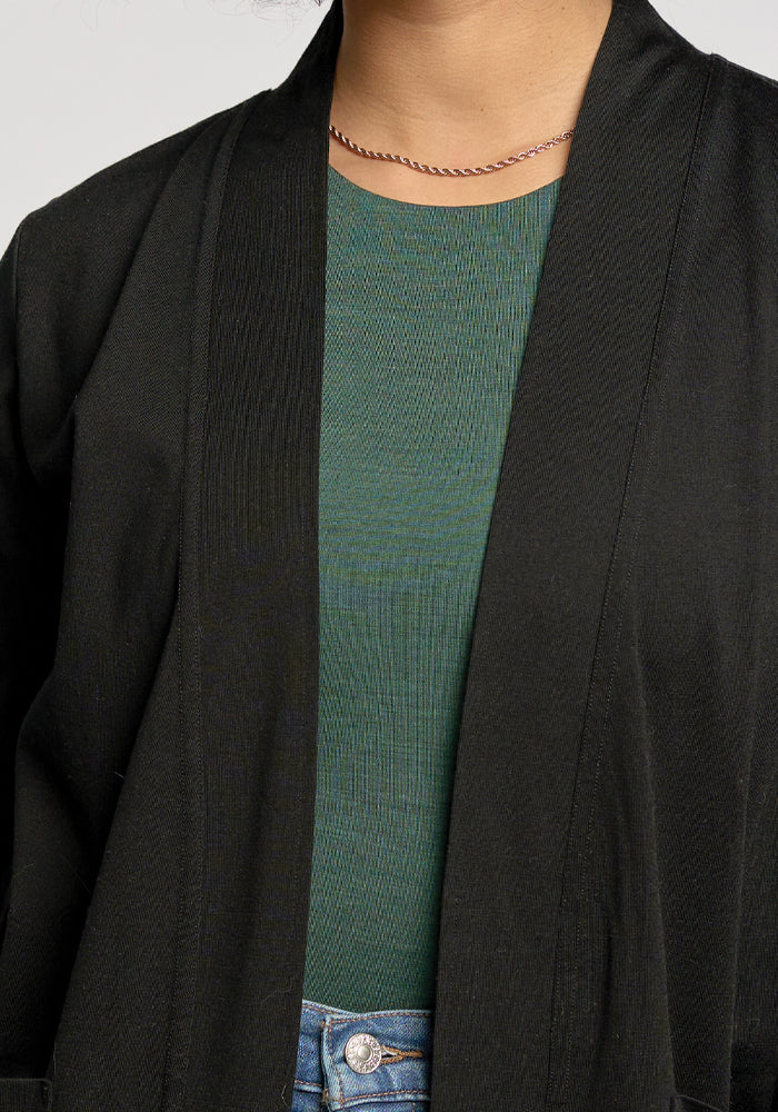 A person in a green ribbed top with a thin gold necklace, partially covered by the Woolx Paisley Cardigan in black, perfectly complements blue jeans and highlights the refined details harmoniously.