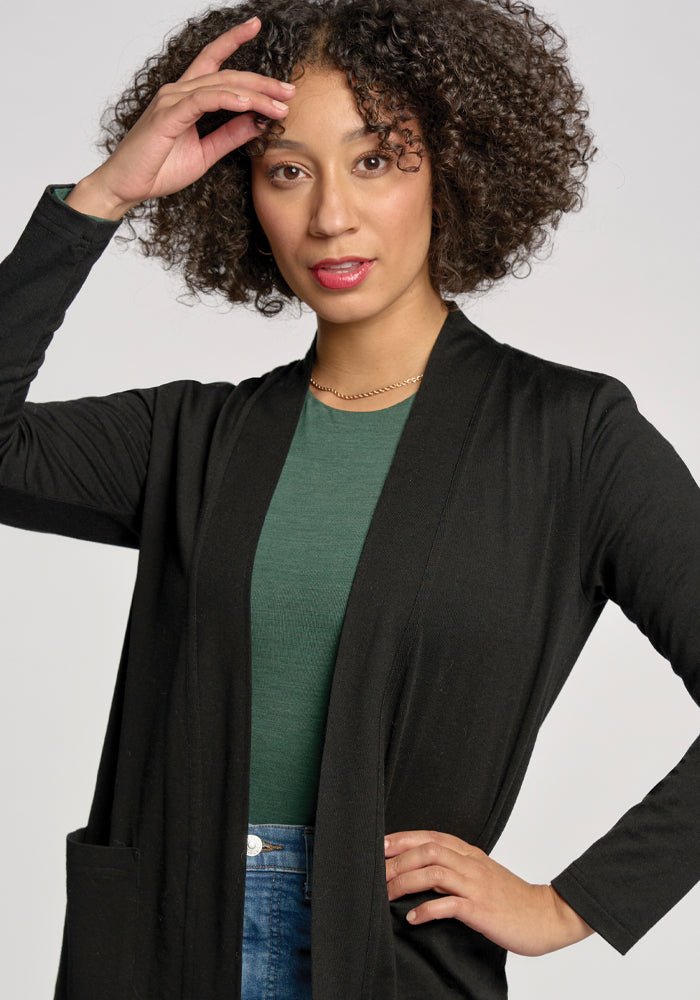 A confident woman with curly hair models against a plain background, wearing a green top under the Woolx Paisley Cardigan in black and jeans. One hand is raised near her forehead, while the other rests on her hip.