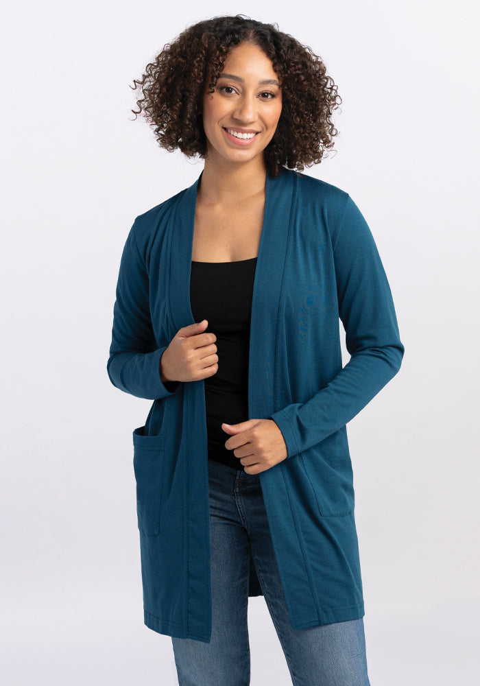 A person with curly hair smiles while wearing the Woolx Paisley Cardigan in Real Teal over a black top and blue jeans. They are standing against a plain white background, with one hand holding the edge of the cardigan made from a Duralite blend. 