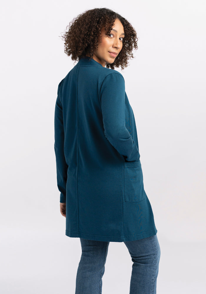 A person with curly hair is standing and looking over their shoulder, wearing the Woolx Paisley Cardigan in Real Teal made from soft Merino wool, paired with jeans against a plain light background.