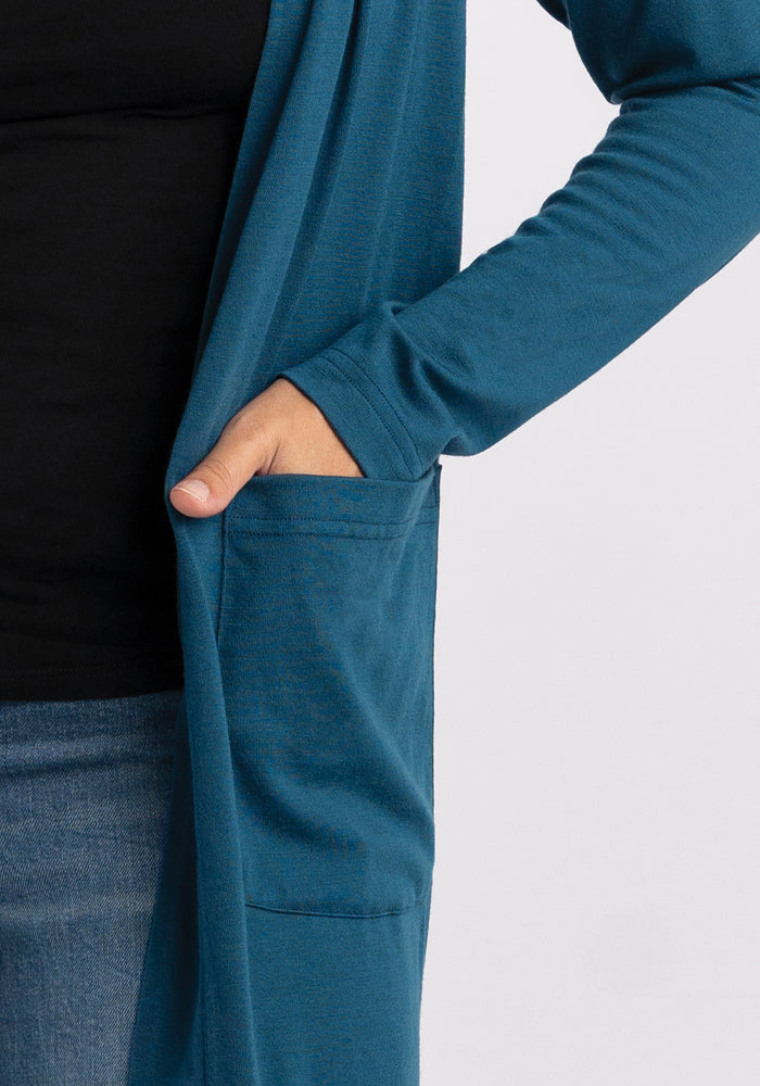 A person is wearing the Woolx Paisley Cardigan - Real Teal, crafted from Australian Merino Wool. Their right hand is tucked into one of its pockets, and the long sleeves complement their black top and denim jeans.