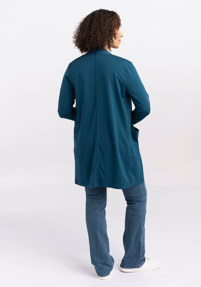 A person with curly hair stands facing away, wrapped in the Paisley Cardigan - Real Teal by Woolx, paired with blue jeans. The backdrop is a clean, plain white.