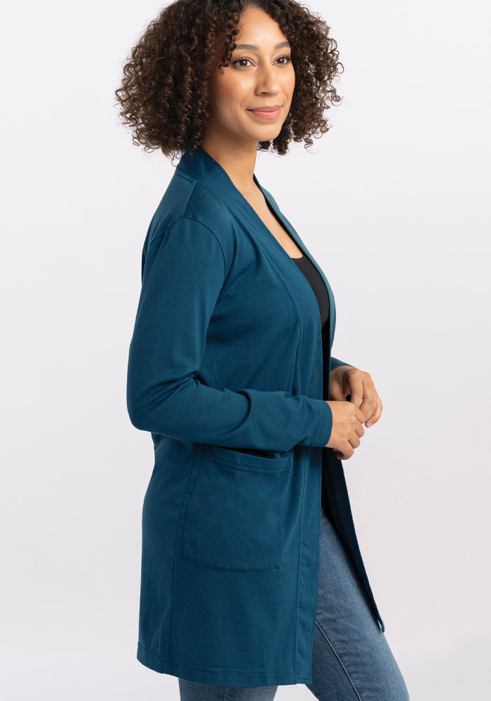 A woman with curly hair smiles while wearing a Woolx Paisley Cardigan - Real Teal over a black top and blue jeans. She stands against a plain white background, highlighting the vibrant pattern.