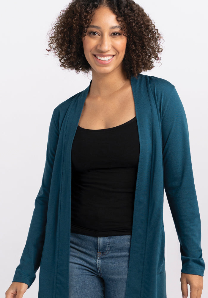 A person with curly hair smiles while wearing the Woolx Paisley Cardigan in Real Teal over a black top and blue jeans, posed against a plain white background.