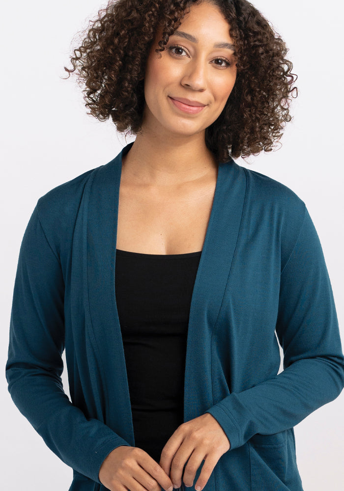 A person with curly hair wearing the "Paisley Cardigan - Real Teal" by Woolx over a black top smiles at the camera against a plain white background.