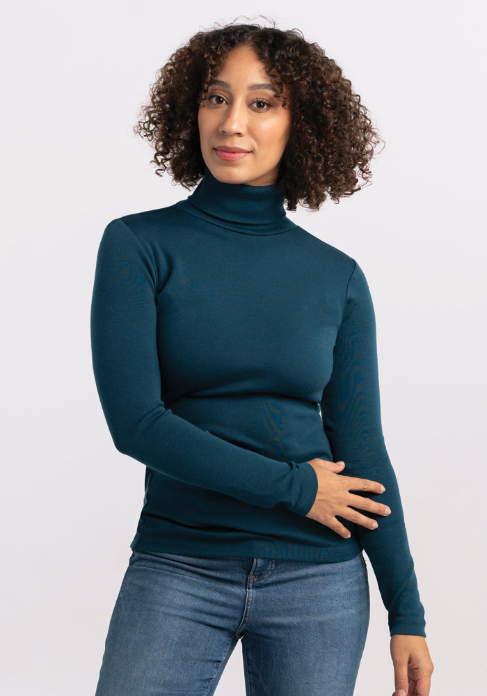 A person with curly hair is wearing the Peyton Turtleneck, a midweight teal Merino wool sweater by Woolx, along with jeans. They are standing against a plain light background, exuding a calm expression as their right hand rests on their left arm. 