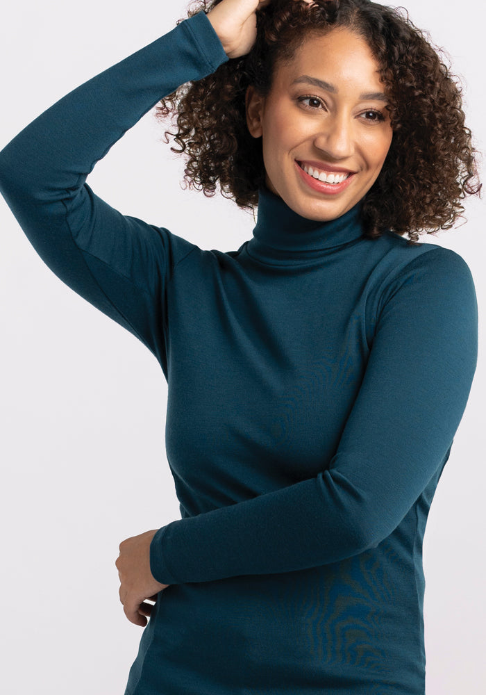 A person with curly hair wearing a midweight teal Peyton Turtleneck by Woolx is smiling and has one arm raised, hand touching their head. The background is plain white.