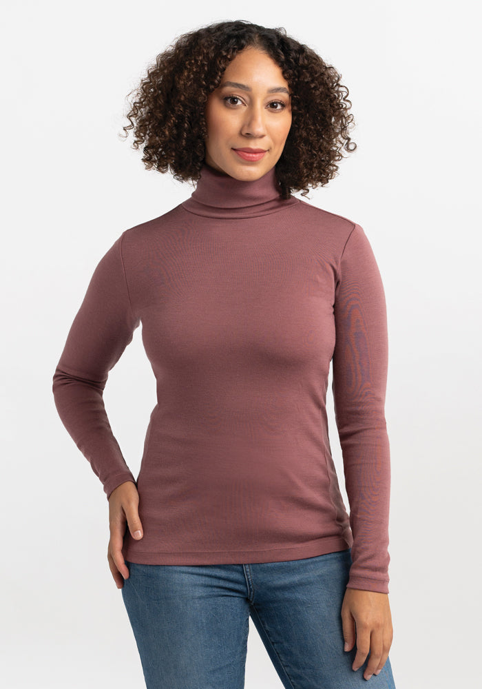 A person with curly hair is standing against a plain white background. They are wearing the Peyton Turtleneck from Woolx in mauve and blue jeans. Both hands are resting on the sides of their hips. 