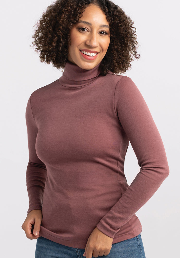 A person with curly hair is smiling and posing in the ultra-soft Peyton Turtleneck from Woolx. They are standing against a plain white background, wearing blue jeans and appearing relaxed with a hand resting on their side.