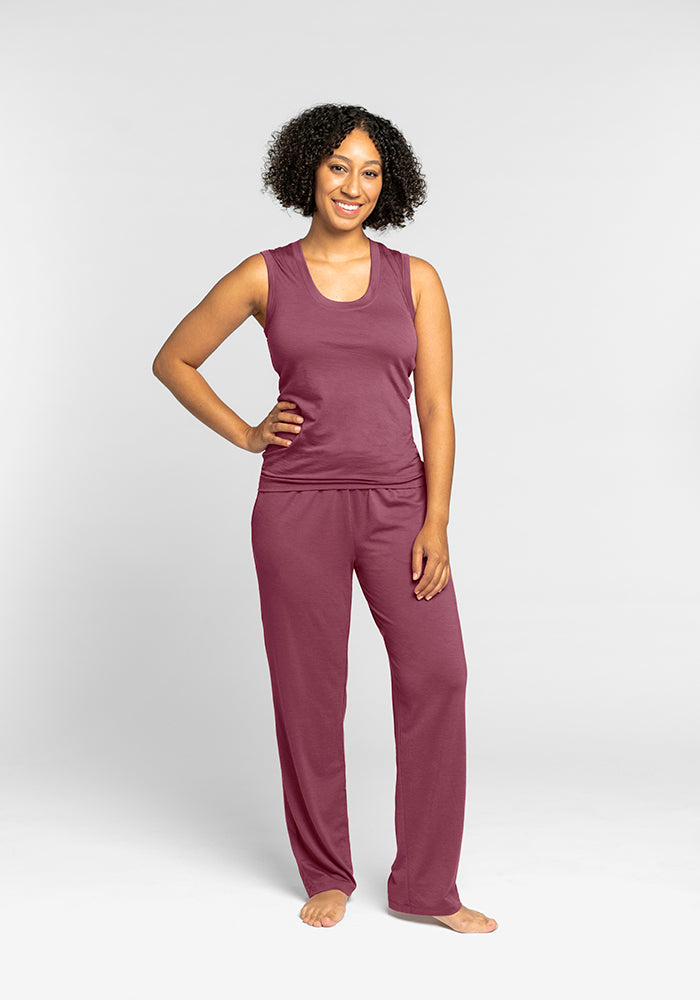 A person stands smiling, wearing the Phoebe Lounge Tank in Wild Ginger and matching pants from Woolx. They have curly hair and are barefoot, with one hand on their hip. The odor-free outfit provides perfect comfort against the plain, light gray background.