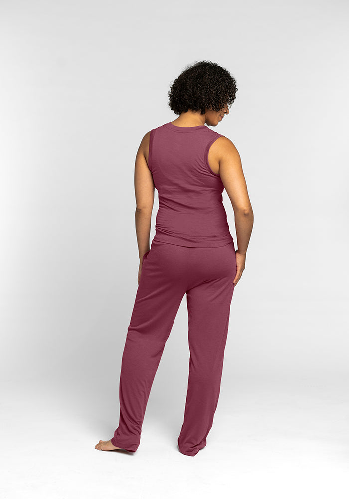 A person with curly hair stands barefoot, facing away from the camera. They're wearing a sleeveless top and matching pants designed for comfort, like the cozy Phoebe Lounge Tank in Wild Ginger from Woolx, against a plain white background.