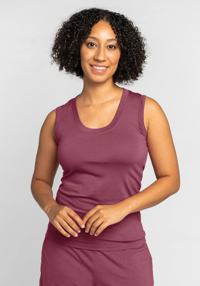 A person with curly hair is smiling, wearing the Phoebe Lounge Tank - Wild Ginger by Woolx, a maroon sleeveless top made of merino wool renowned for its cooling comfort, against a plain background. 