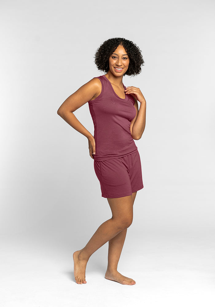 A person with curly hair is smiling and posing in a sleeveless Phoebe Lounge Tank in Wild Ginger by Woolx. They are standing barefoot on a plain white background, radiating comfort in an odor-free ensemble.