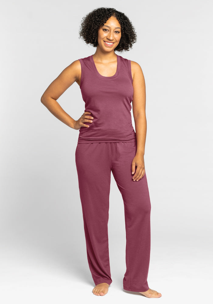 A person with curly hair stands smiling, wearing the Phoebe Lounge Tank in Wild Ginger from Woolx, paired with matching loose pants that resemble breathable pajamas. They are barefoot, standing against a plain white background.