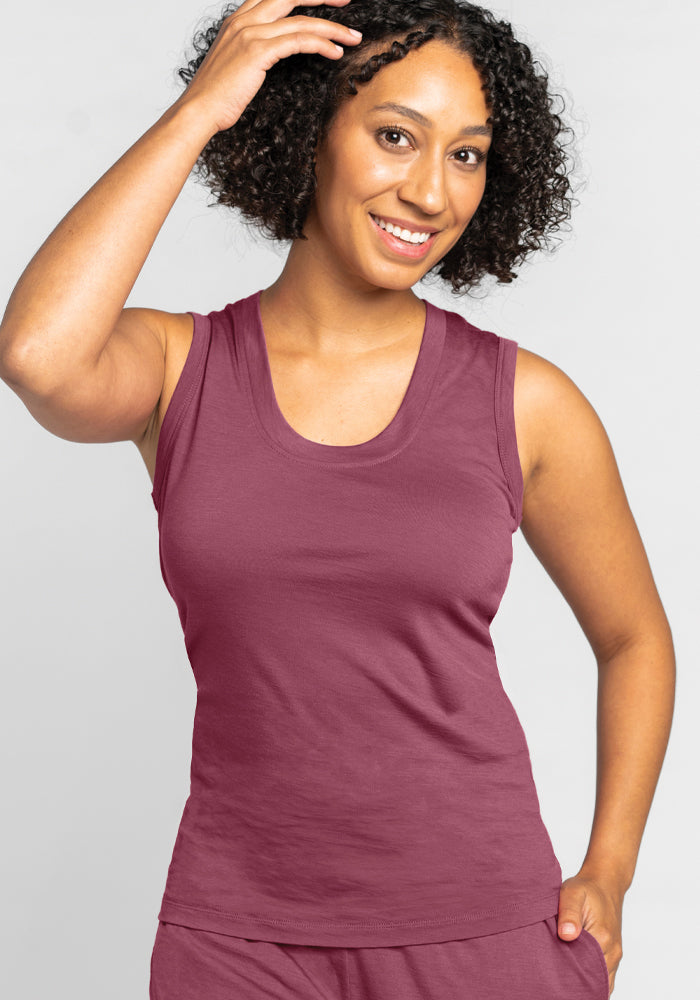 A smiling person with curly hair, dressed in the Phoebe Lounge Tank in Wild Ginger by Woolx, paired with matching pants, stands against a plain background with one hand touching their hair.