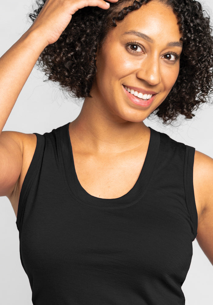 A person with curly hair smiles at the camera, dressed in the Phoebe Lounge Tank - Black by Woolx, a sleeveless top crafted from lightweight Merino wool, with one hand gently touching their hair. The background is a light gray color.