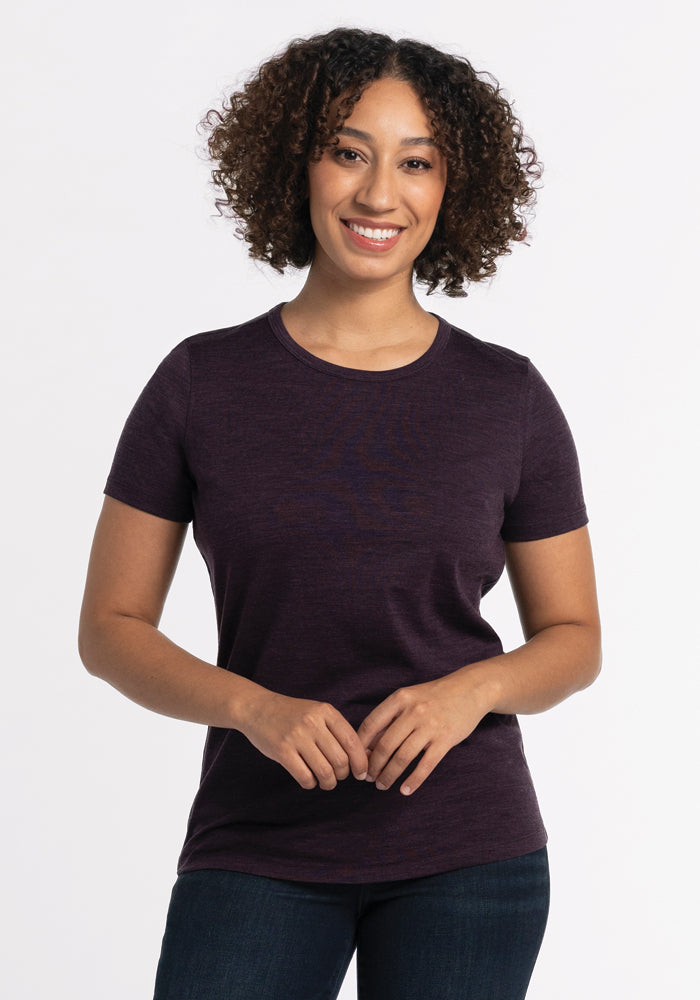 A person with curly hair wearing the Woolx Liza Crew Neck Tee in Deep Plum and blue jeans stands against a white background, smiling. They have their hands clasped in front of them. 