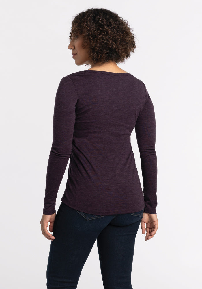 A person with curly hair is standing and facing away from the camera, wearing a long-sleeved Layla V Neck Top in deep plum by Woolx and dark jeans. The background is plain and white.