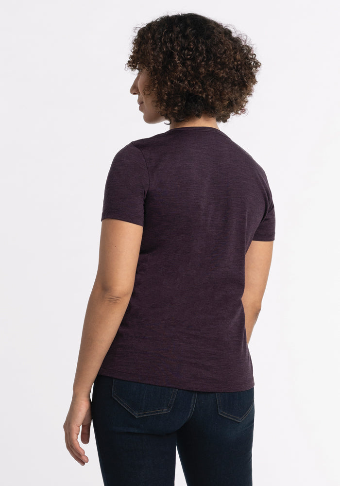 A person with curly hair faces away from the camera, wearing a Woolx Liza Crew Neck Tee in Deep Plum and dark jeans, set against a simple light background.