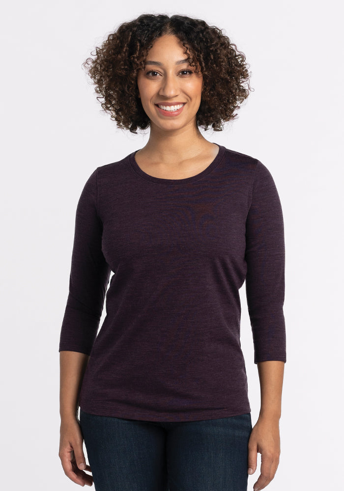 A person with curly hair smiles while standing against a white background, wearing the Woolx Jenny 3/4 Sleeve Crew Neck in Deep Plum paired with dark jeans. 