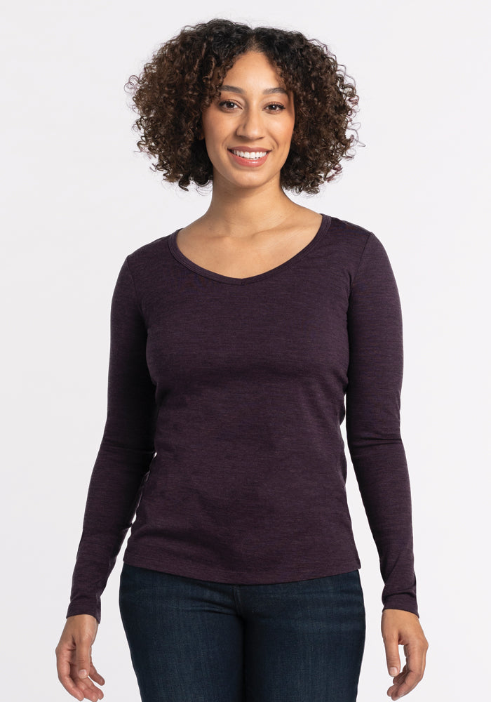 A person with curly hair smiles while wearing the Woolx Layla V Neck Top in Deep Plum and jeans, standing against a plain white background.