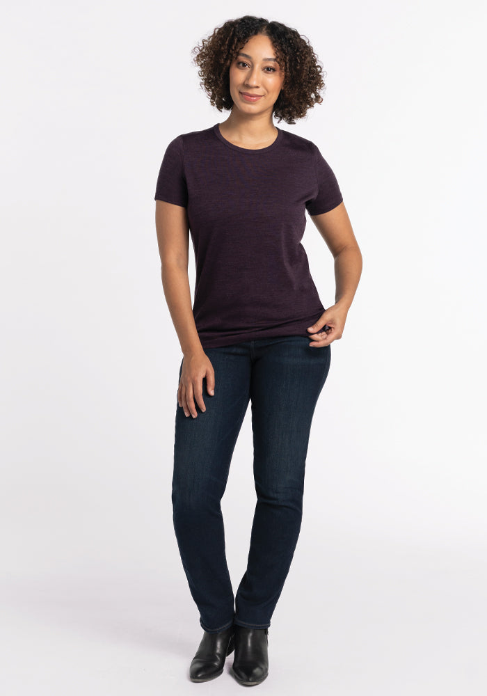 A person with curly hair stands against a plain white background, wearing the Liza Crew Neck Tee in Deep Plum by Woolx, accompanied by dark jeans and black ankle boots. They are smiling gently, with their hands resting casually.