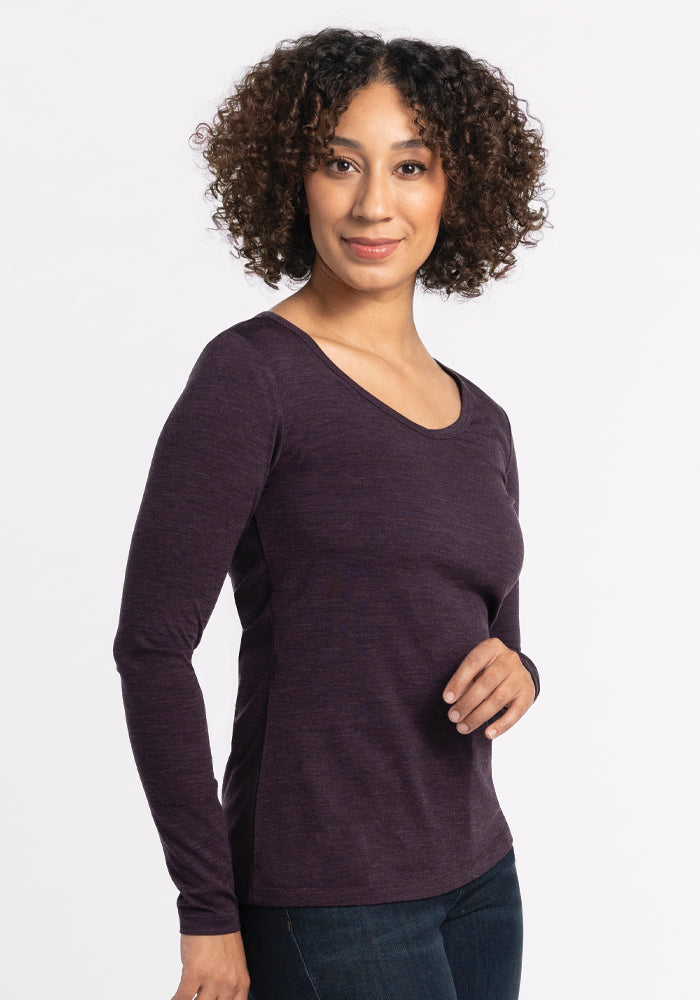 A person with curly hair smiles while wearing a long-sleeve Layla V Neck Top in Deep Plum by Woolx and jeans. They are standing against a plain white background. 