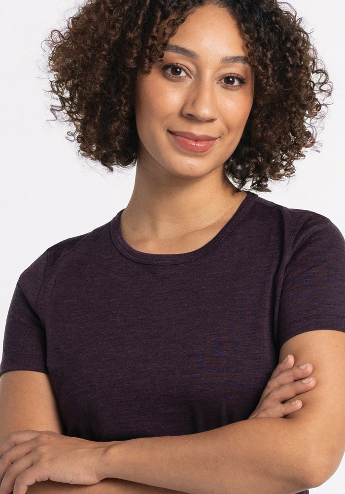 A person with curly hair wearing the Liza Crew Neck Tee in Deep Plum by Woolx, made of lightweight fabric, stands with their arms crossed, smiling gently against a plain white background.