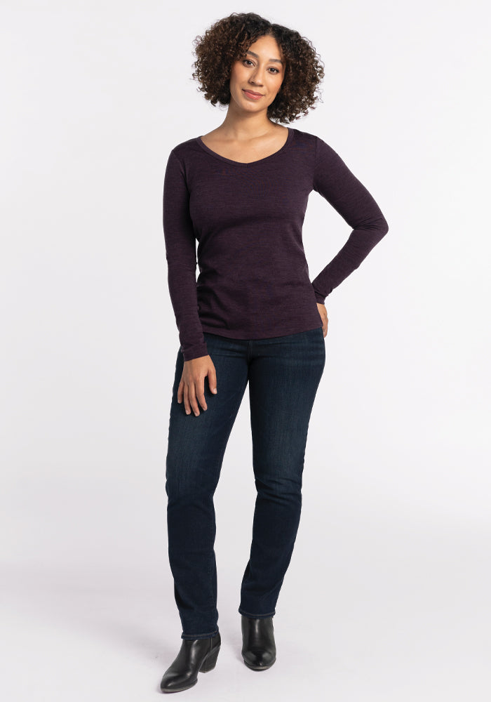 A person with curly hair is wearing a deep plum Layla V Neck Top from Woolx and blue jeans, standing with one hand on their hip. They are also wearing black ankle boots against a plain white background.