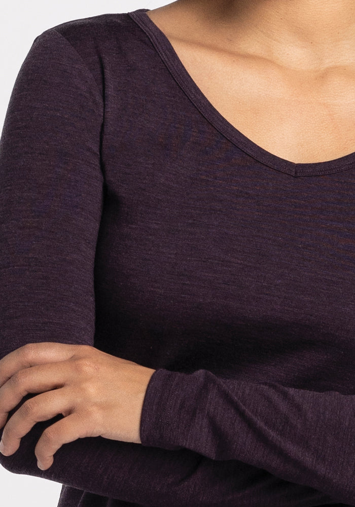 A person wearing a deep plum Layla V Neck Top by Woolx crosses their arms. This Merino Wool performance top features a stylish V-neck design against a plain white background.